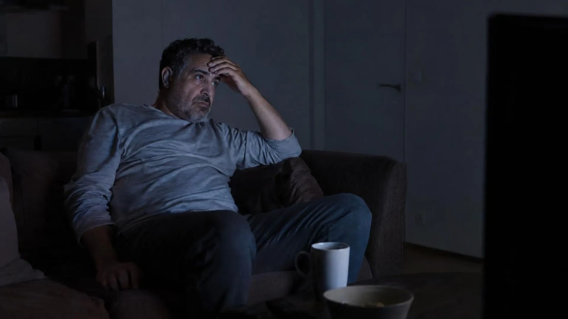 Iemand zit in een verder donkere kamer met alleen het schijnsel van een tv, een hand tegen het voorhoofd