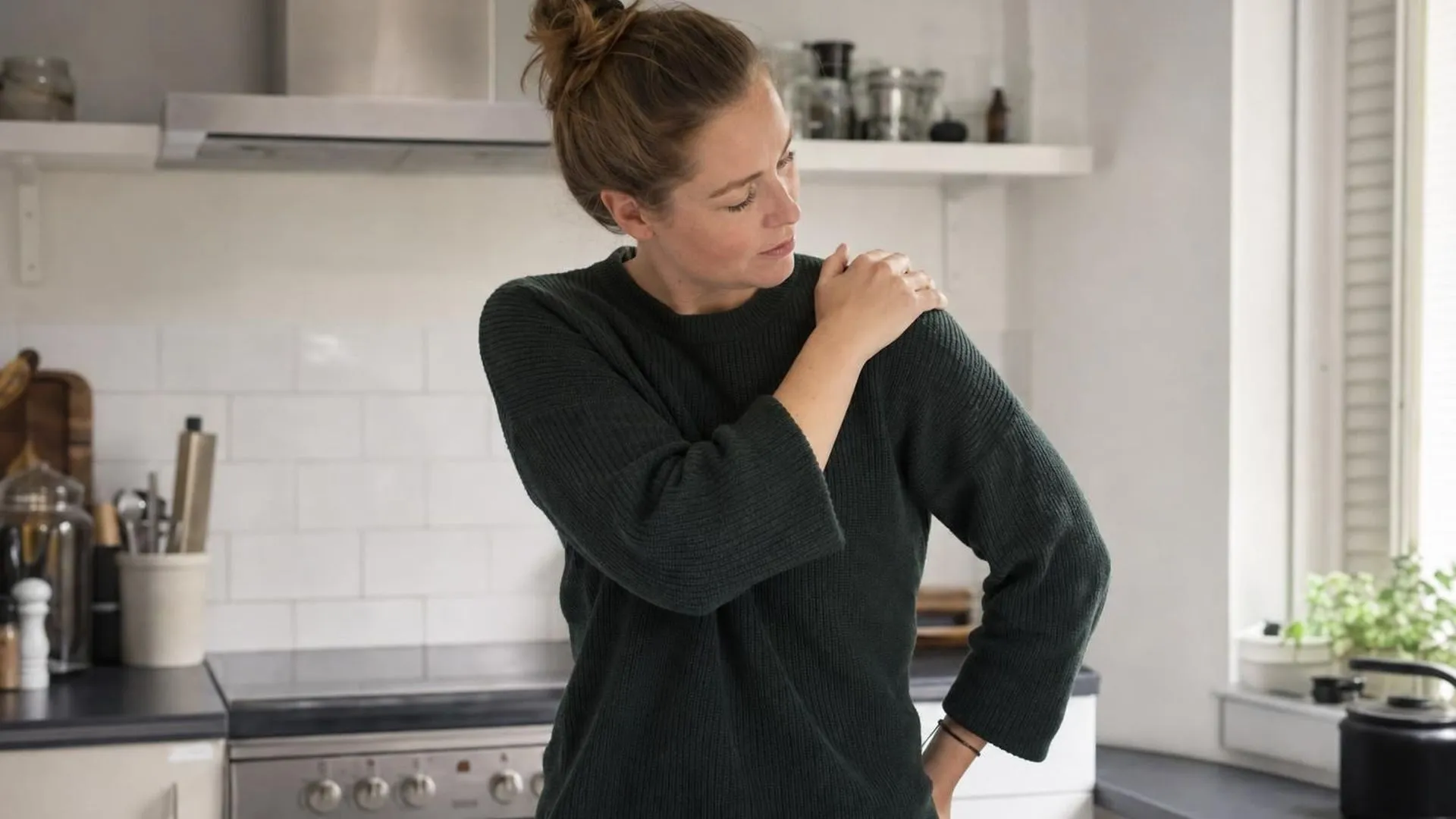 Iemand wrijft 's ochtends over een stijve schouder in een rustige keuken