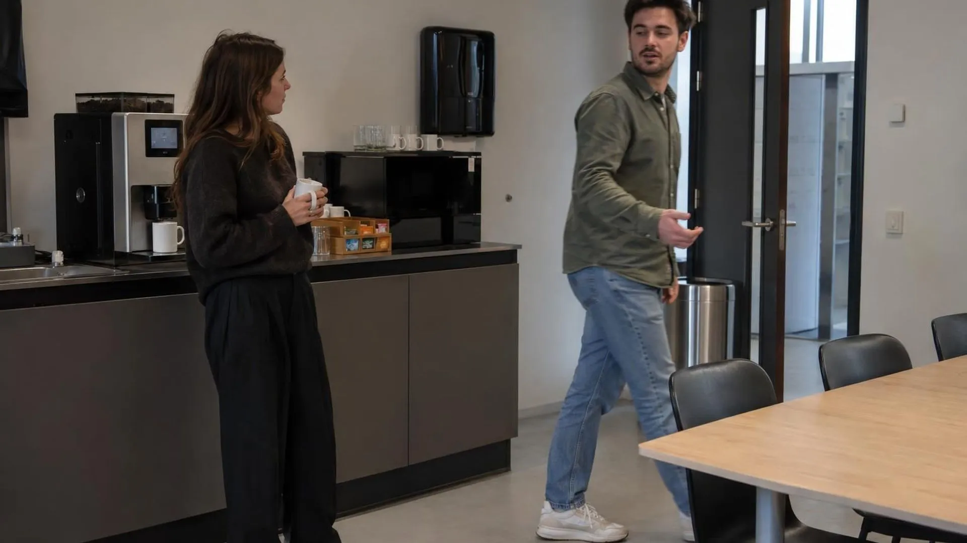 Twee collega's in een pauzeruimte, de een loopt al weg midden in een zin, de ander blijft alleen achter bij het koffieapparaat.