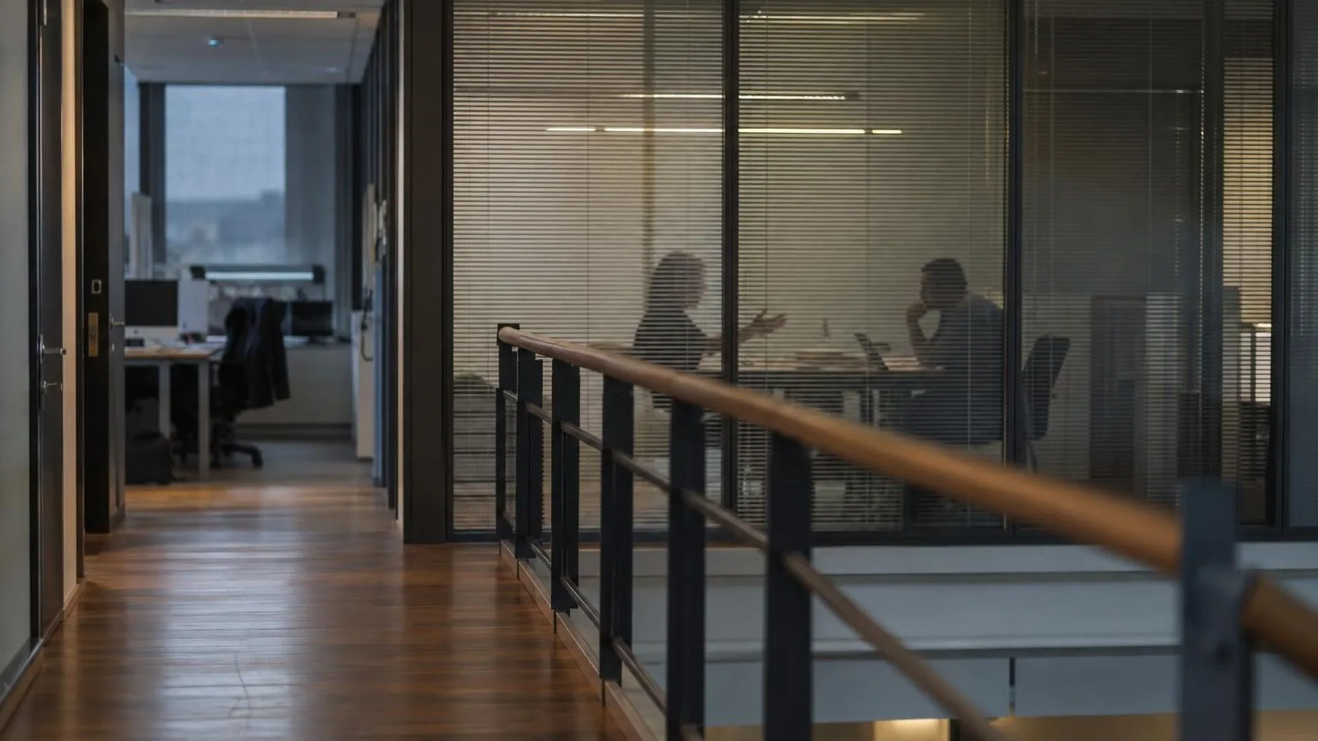 Door een glazen wand met half dichte jaloezieën is een gesprek in een vergaderkamer te zien, wazig op de achtergrond