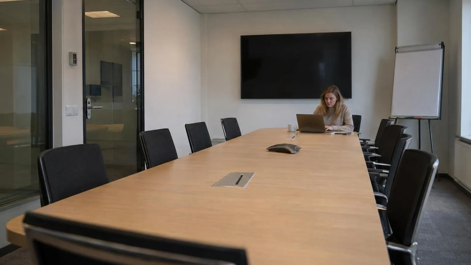 Eén persoon zit alleen aan het uiteinde van een lange vergadertafel met verder alleen lege stoelen, in kil kantoorlicht