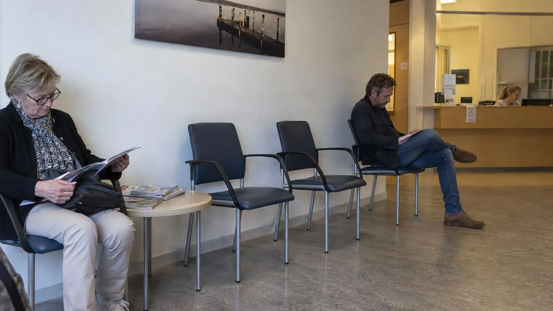 Een rij lege wachtkamerstoelen naast een tafeltje met tijdschriften, in vaal tl-licht bij de huisarts.