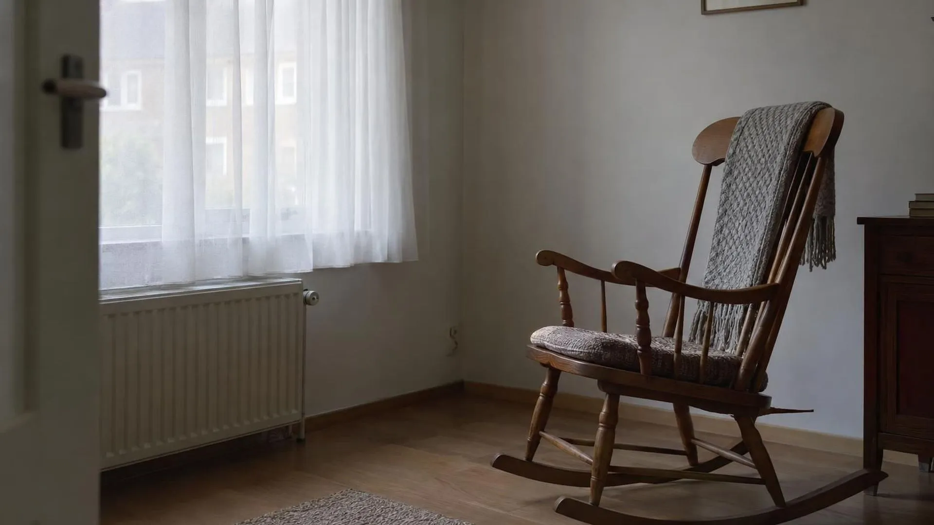 Een lege schommelstoel in een stille, sober ingerichte kamer met daglicht door een vitrage