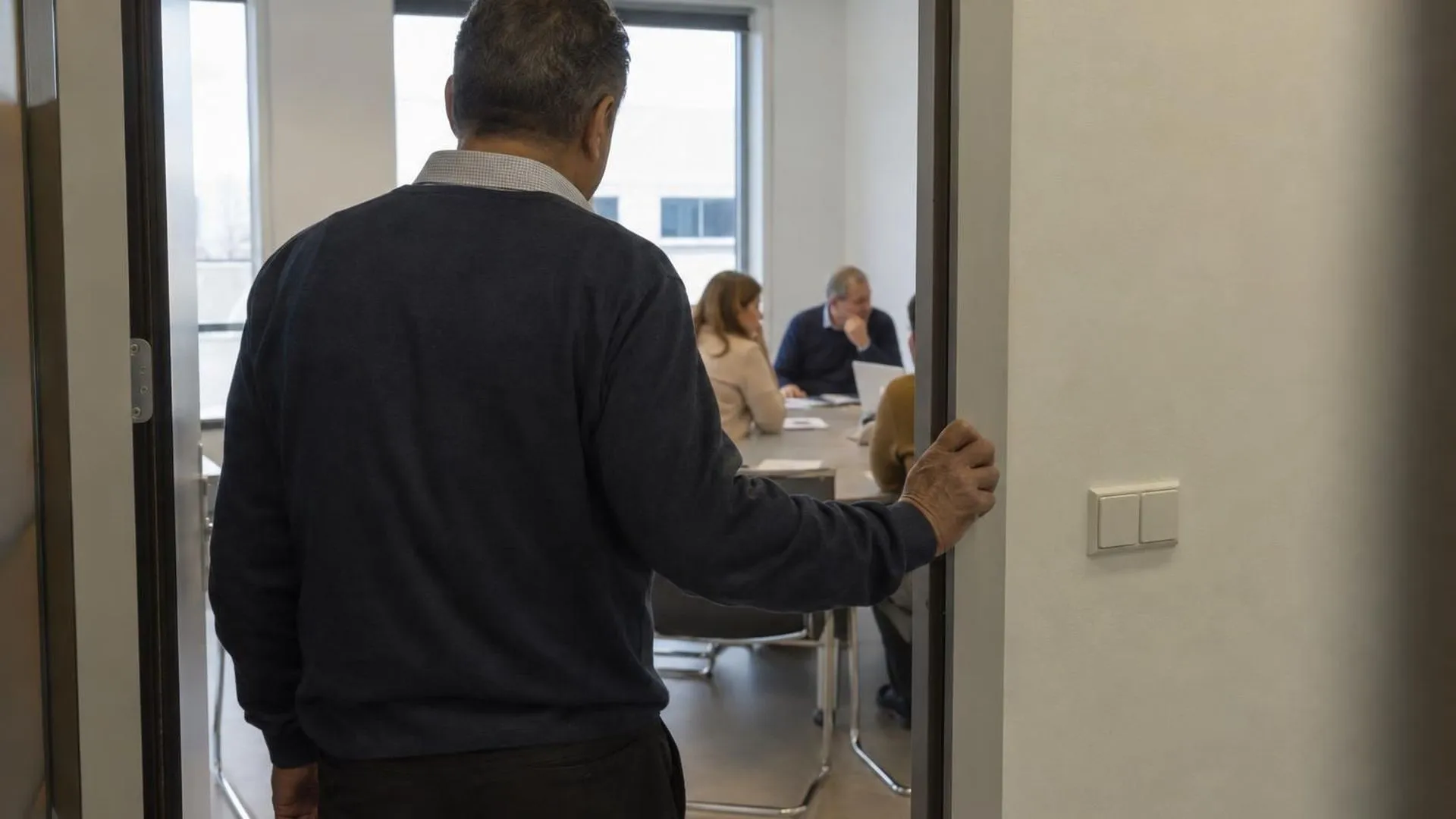 Van achteren gezien blijft iemand met een hand op de deurpost staan, aarzelend voor het een verlichte vergaderruimte inloopt.