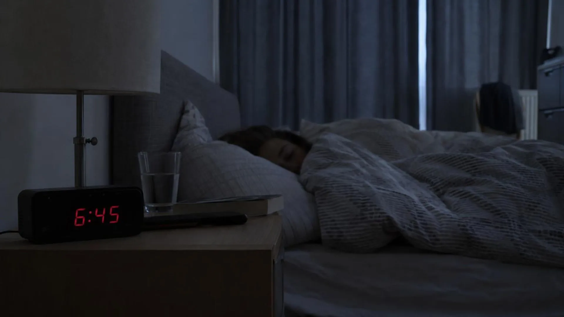 Een wekker die 6:45 aangeeft op het nachtkastje, de gordijnen zijn nog dicht