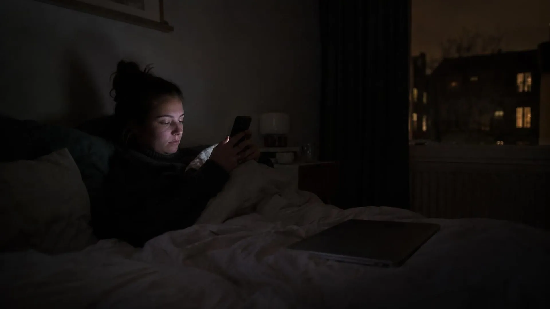Een telefoonscherm dat oplicht in een donkere kamer, een dichte laptop ligt er onaangeraakt naast