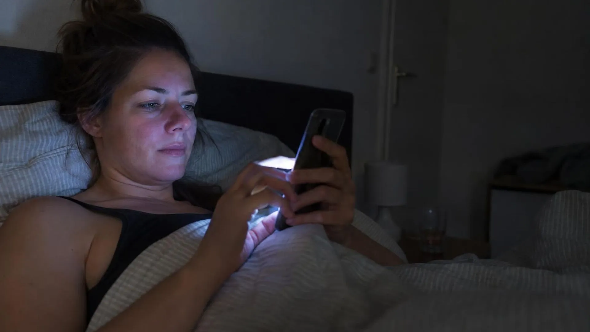 Handen die scrollen op een telefoon in bed, met blauw schermlicht in een verder donkere slaapkamer.