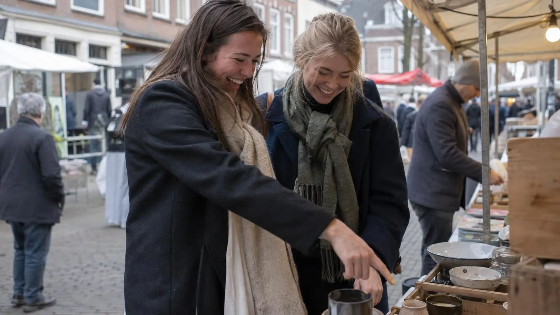 Twee vrienden struinen samen over een zaterdagse marktkraam, een van hen wijst lachend naar iets.