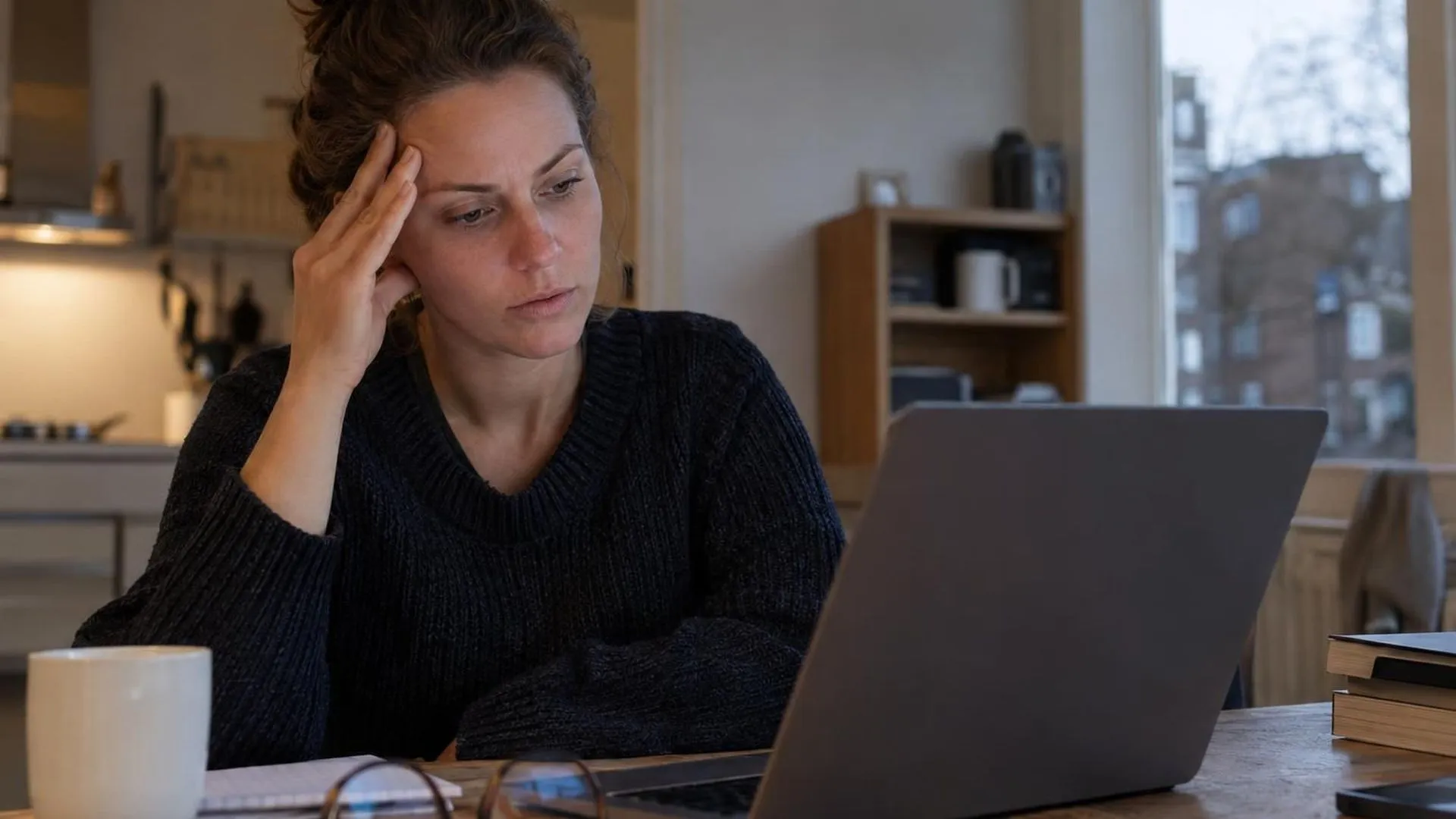 Close-up van een hand tegen een slaap gedrukt, met een laptop weerspiegeld in een bril op het bureau