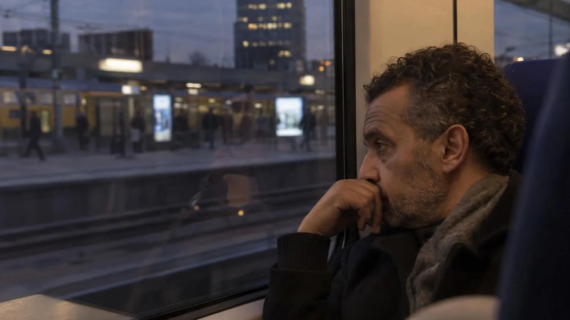 Een vage reflectie van iemand in een treinraam bij schemer, met voorbijglijdende lichtjes en perronborden op de achtergrond.