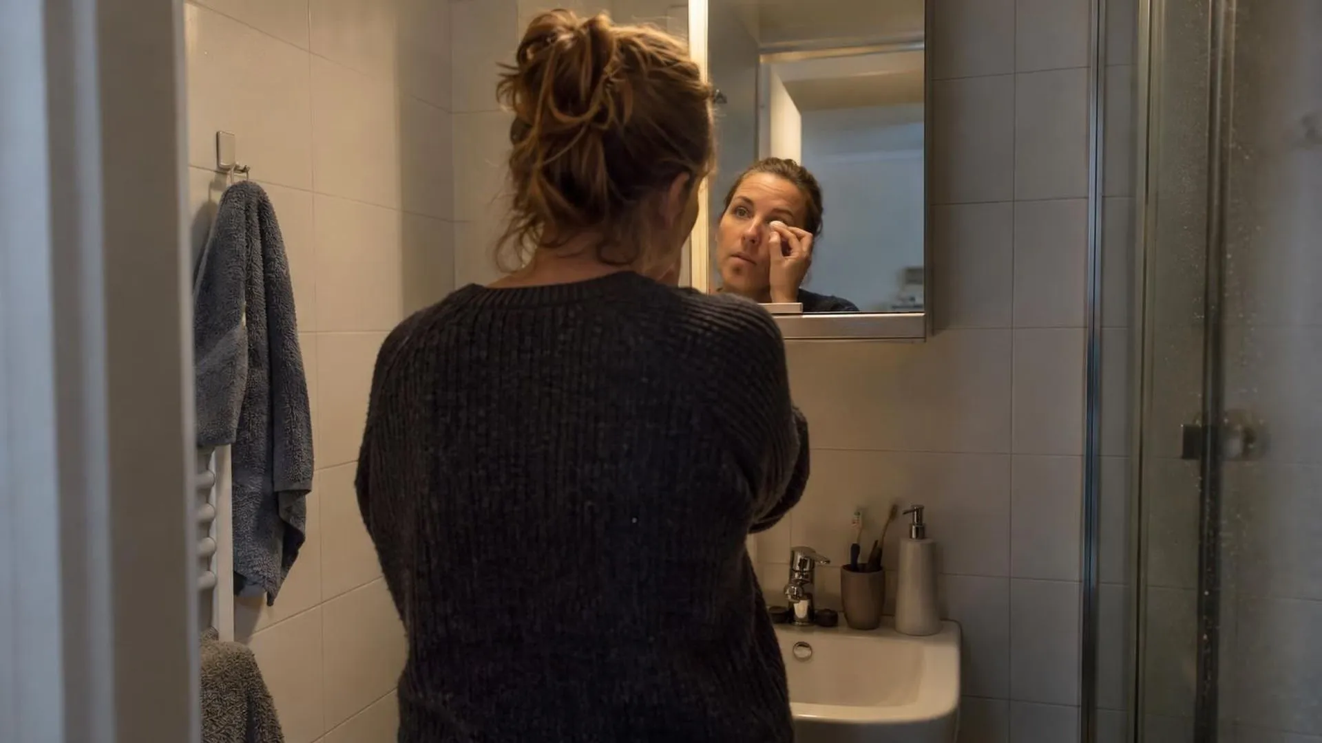 Gezien van achteren: iemand kijkt in een badkamerspiegel bij kaal lamplicht op een doordeweekse ochtend.