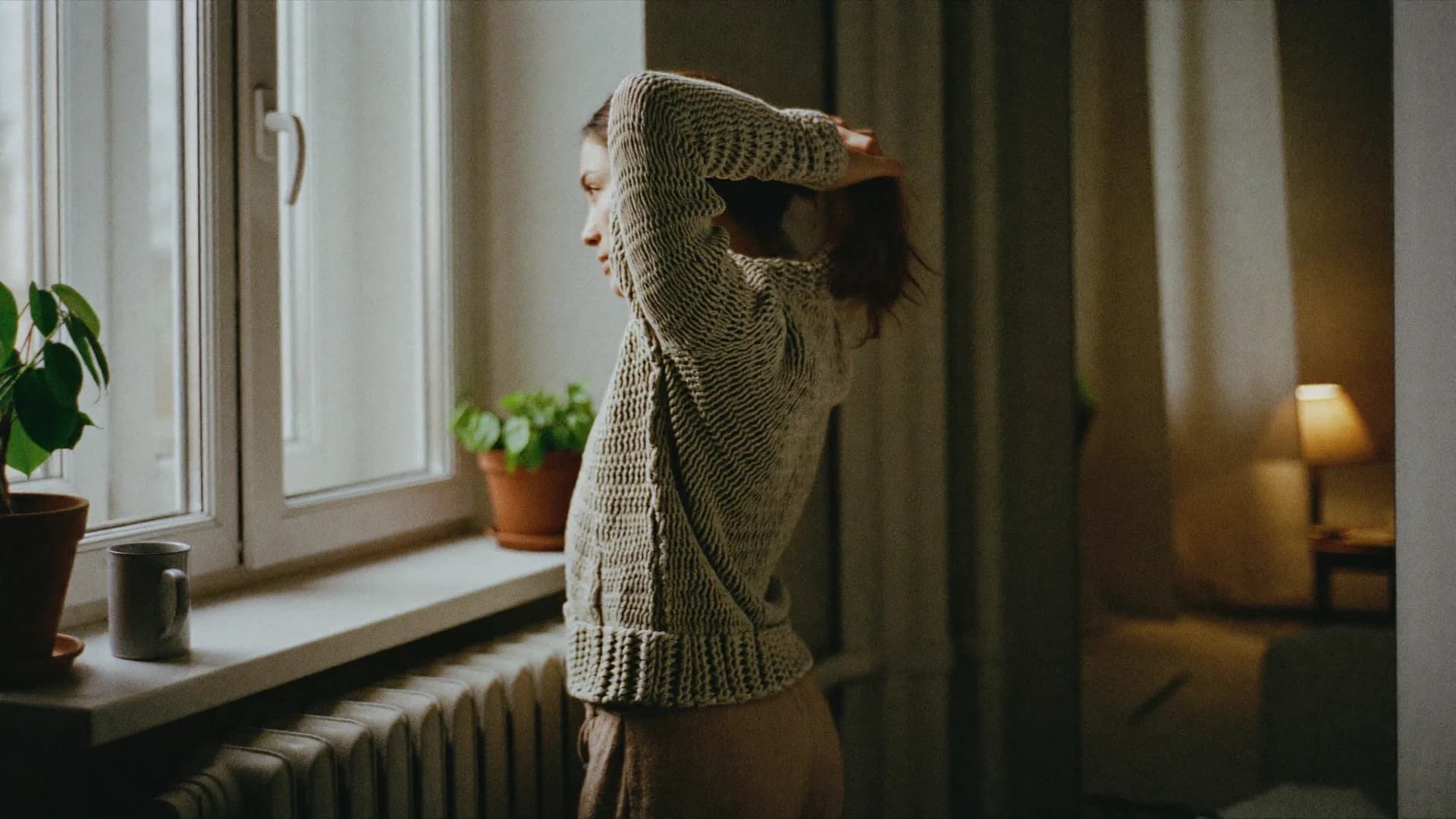 Vrouw in een trui bij het raam die haar haar opsteekt in het ochtendlicht