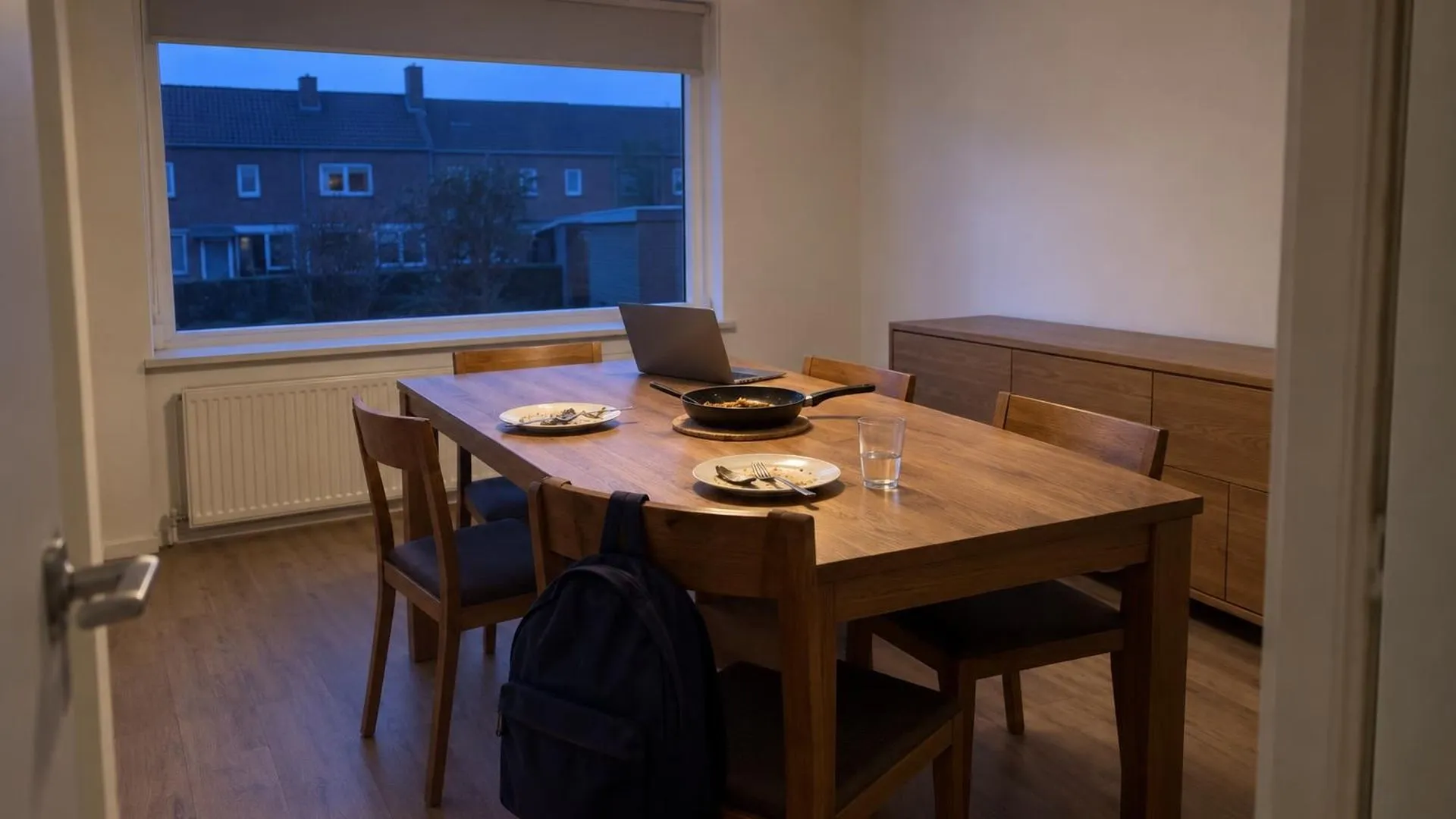Een opengeklapte werklaptop tussen de vuile borden op een eettafel na het zondagse avondeten, gezien vanuit de deuropening in de schemering