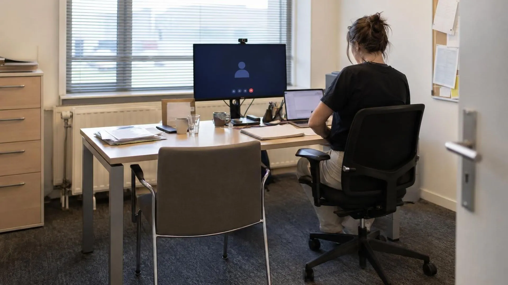 Een lege bezoekersstoel bij een bureau, met op de achtergrondmonitor een onbeantwoorde video-oproep.