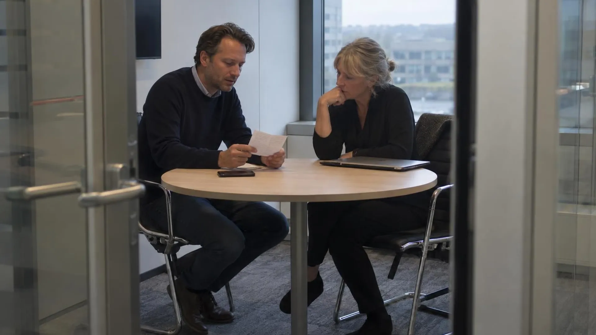 In een kleine vergaderruimte leest een man voor van een handgeschreven briefje, een collega luistert aandachtig.