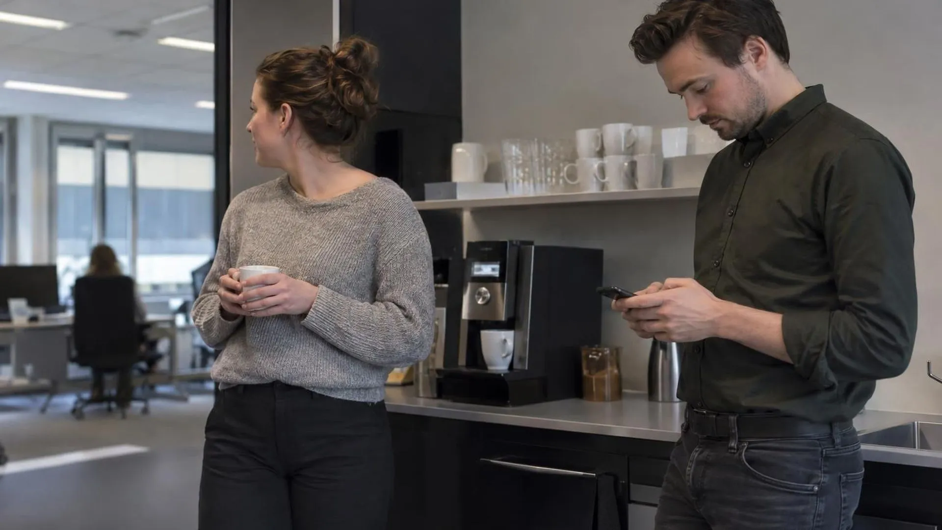 Twee collega's bij het koffiehoekje op kantoor, waarbij de een zich afwendt terwijl de ander op een telefoon kijkt