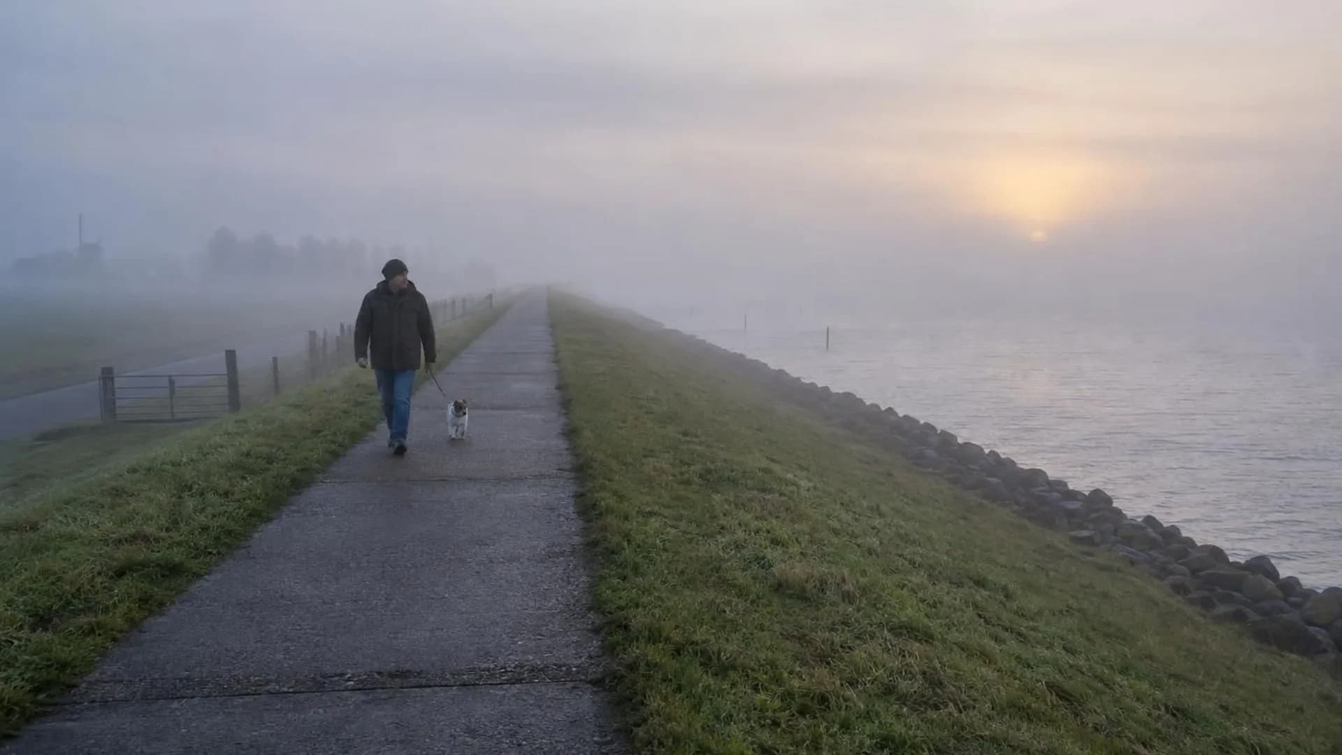 Iemand loopt klein over een lange dijk in de vroege ochtendmist