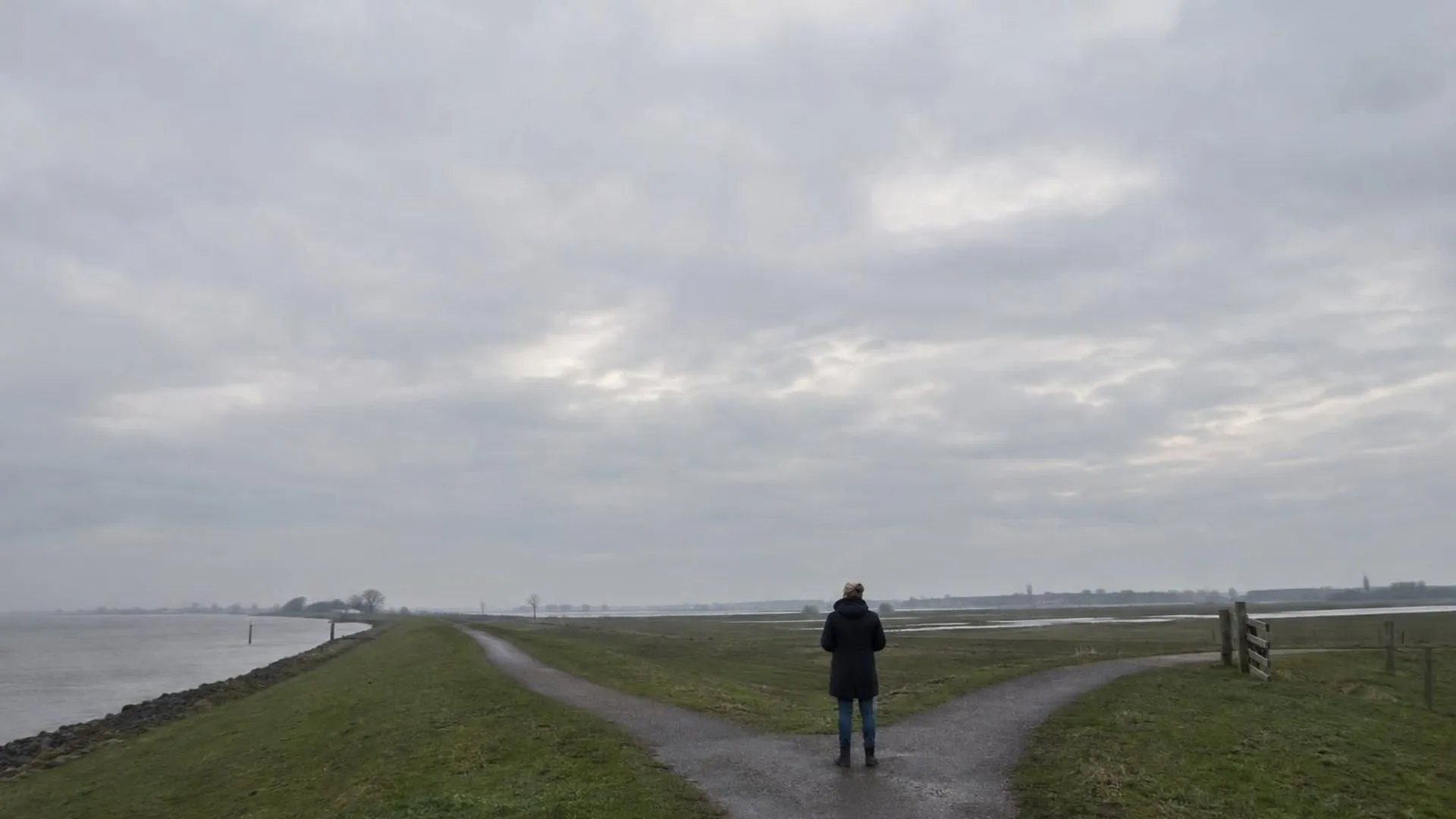 Een klein figuur bij een splitsing op een dijkpad, met een groot grijs luchtdeel in beeld